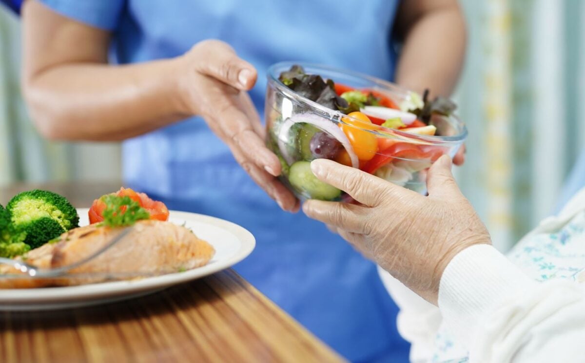 Photo shows a healthcare worker in blue scrubs handing a large glass bowl of salad to an elderly patient