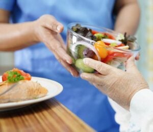Photo shows a healthcare worker in blue scrubs handing a large glass bowl of salad to an elderly patient