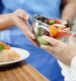 Photo shows a healthcare worker in blue scrubs handing a large glass bowl of salad to an elderly patient