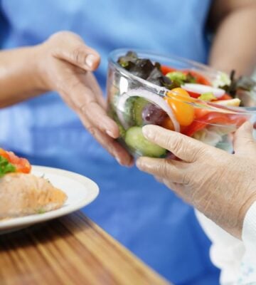 Photo shows a healthcare worker in blue scrubs handing a large glass bowl of salad to an elderly patient