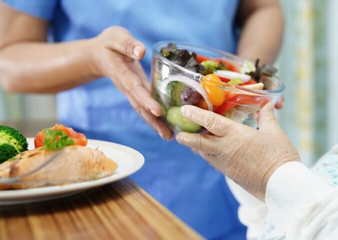 Photo shows a healthcare worker in blue scrubs handing a large glass bowl of salad to an elderly patient