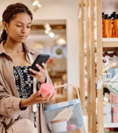 Photo shows a young woman looking up the ingredients of a beauty product on her phone in a store