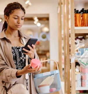 Photo shows a young woman looking up the ingredients of a beauty product on her phone in a store