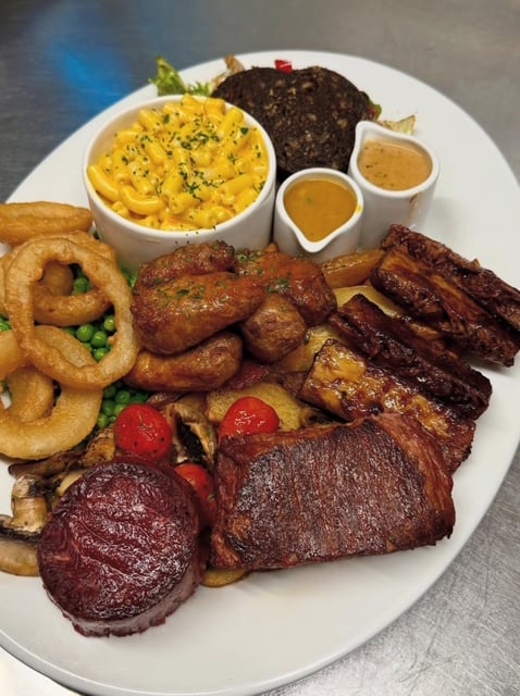 Photo shows a platter of grilled plant-based meat, onion rings, mac and cheese, and other sides from the vegan steakhouse The Queen Inn