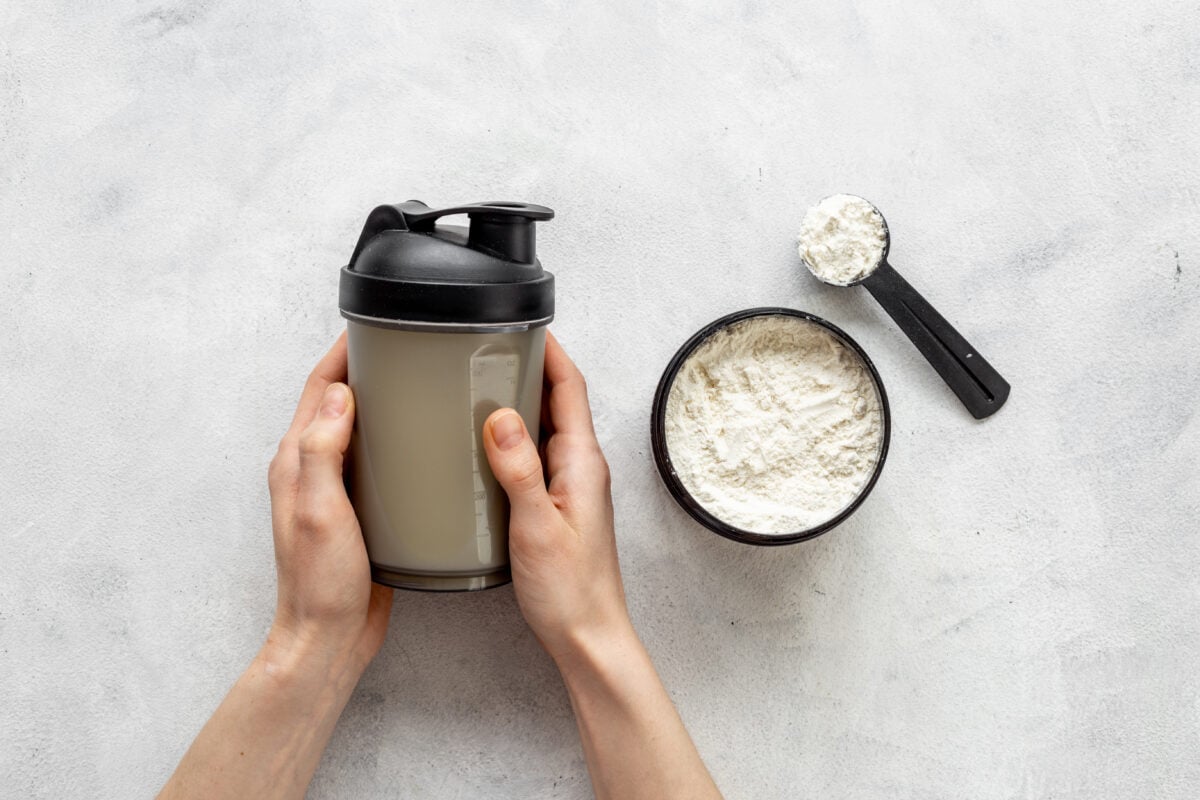 Photo shows someone's hands as they hold a protein shaker next to a tub and scoop of protein powder