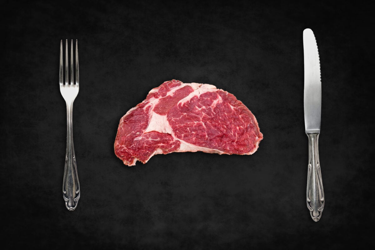 Photo shows a raw steak on a black tablecloth flanked by a silver knife and fork