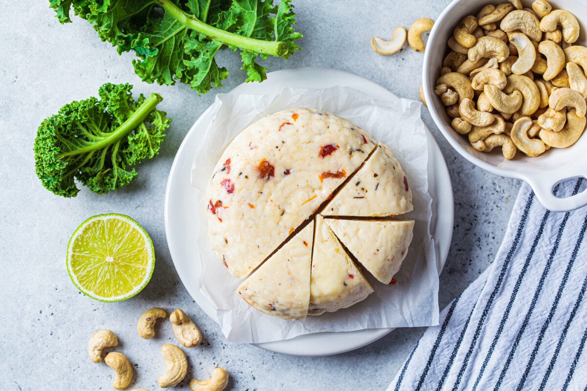 Photo shows a wheel of plant-based cheese alongside half a lemon, some greens, and a dish of cashew nuts