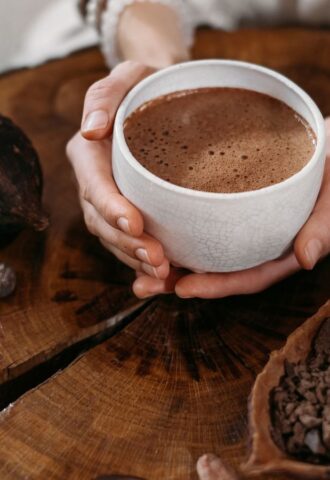 Photo shows a woman's hands holding a cup of hot chocolate, next to a large cocoa pod and a wooden bowl of cocoa beans