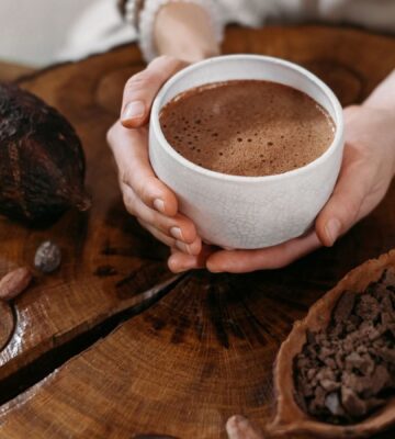 Photo shows a woman's hands holding a cup of hot chocolate, next to a large cocoa pod and a wooden bowl of cocoa beans