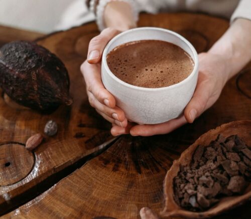 Photo shows a woman's hands holding a cup of hot chocolate, next to a large cocoa pod and a wooden bowl of cocoa beans