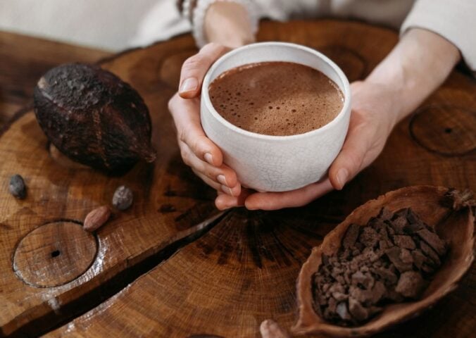 Photo shows a woman's hands holding a cup of hot chocolate, next to a large cocoa pod and a wooden bowl of cocoa beans