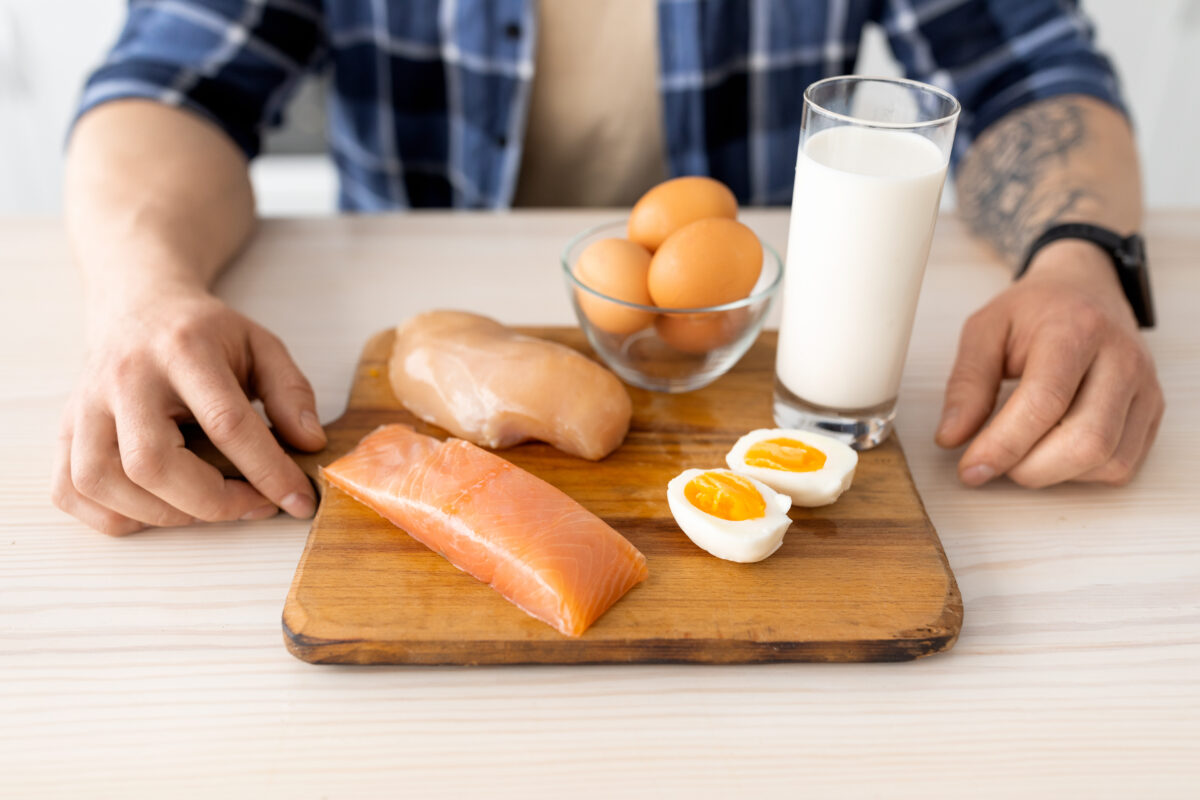 Photo shows a man sat in front of a wood board loaded with animal products, including raw and cooked eggs, a glass of milk, raw fish, and raw poultry