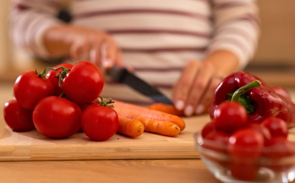 Photo shows a woman chopping carrots, tomatoes, and red peppers on a wooden board