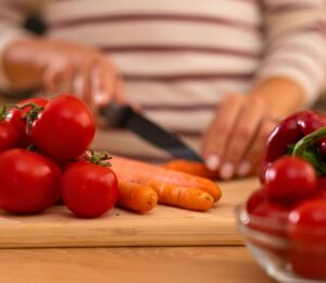 Photo shows a woman chopping carrots, tomatoes, and red peppers on a wooden board