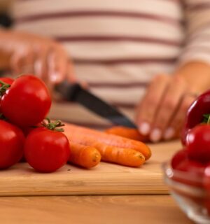 Photo shows a woman chopping carrots, tomatoes, and red peppers on a wooden board