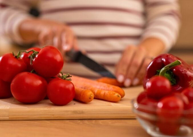 Photo shows a woman chopping carrots, tomatoes, and red peppers on a wooden board