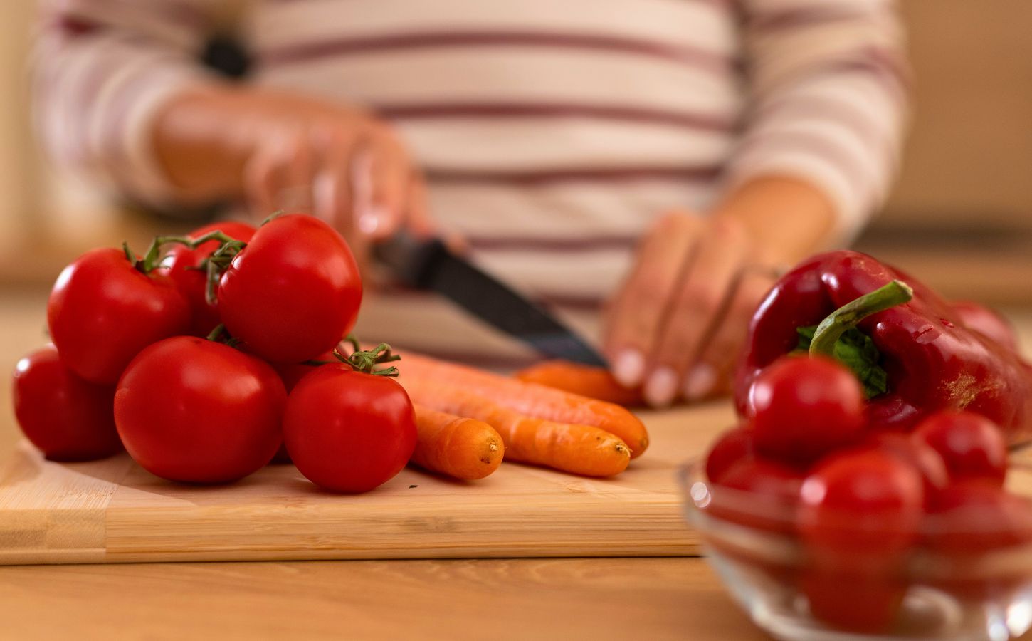 Photo shows a woman chopping carrots, tomatoes, and red peppers on a wooden board