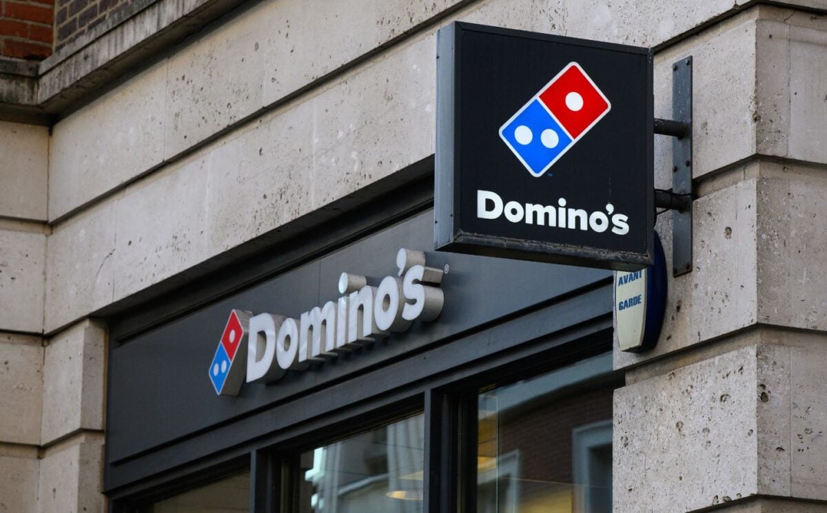 Photo shows the front of a Domino's storefront in the UK, including a projecting sign and a horizontal sign bearing the brand's name and red-and-blue logo