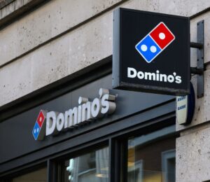 Photo shows the front of a Domino's storefront in the UK, including a projecting sign and a horizontal sign bearing the brand's name and red-and-blue logo