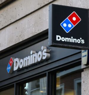 Photo shows the front of a Domino's storefront in the UK, including a projecting sign and a horizontal sign bearing the brand's name and red-and-blue logo