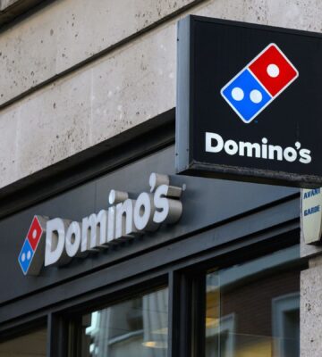 Photo shows the front of a Domino's storefront in the UK, including a projecting sign and a horizontal sign bearing the brand's name and red-and-blue logo