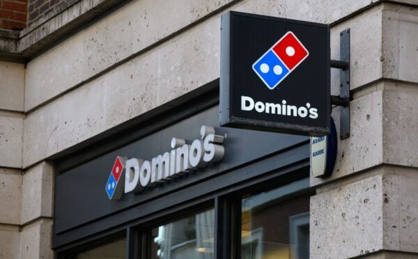Photo shows the front of a Domino's storefront in the UK, including a projecting sign and a horizontal sign bearing the brand's name and red-and-blue logo