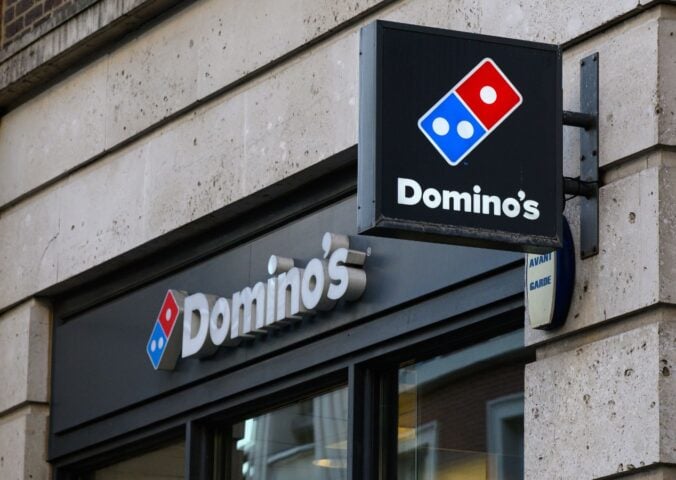 Photo shows the front of a Domino's storefront in the UK, including a projecting sign and a horizontal sign bearing the brand's name and red-and-blue logo
