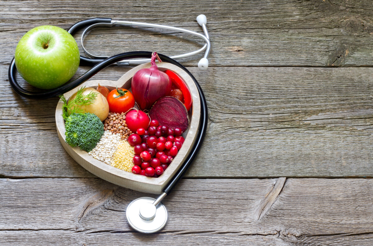 Photo shows a heart-shaped plate full of nutritious plant-based foods like broccoli, berries, tomatoes, and beans, next to a Dr's stethoscope with an apple in it