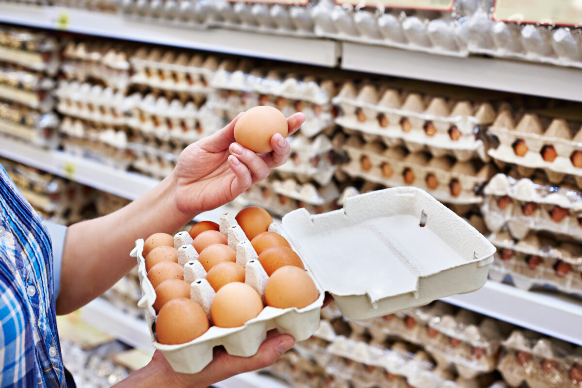 Photo shows a woman holding a large box of eggs in a supermarket, with dozens of similar boxes on shelves in the background