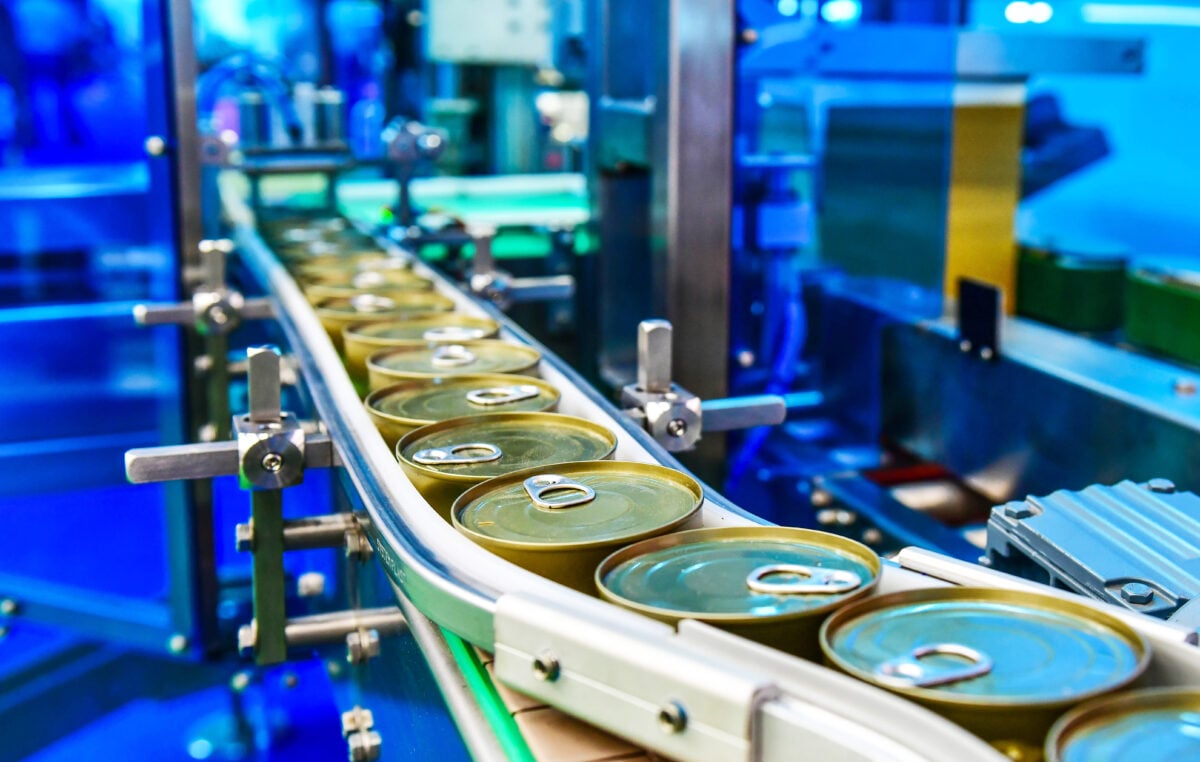 Photo shows a food processing facility and a row of cans on a conveyor belt
