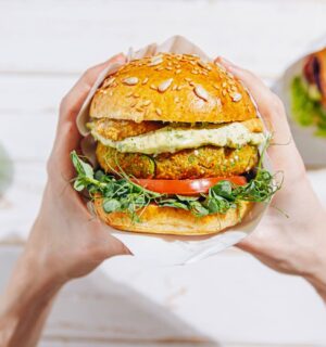 Photo shows a woman's hands holding a large vegetarian burger next to a green smoothie and a second sandwich. A new report suggests that ultra-processed plant-based foods are better for heart health than unprocessed animal foods like meat and dairy