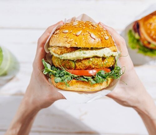 Photo shows a woman's hands holding a large vegetarian burger next to a green smoothie and a second sandwich. A new report suggests that ultra-processed plant-based foods are better for heart health than unprocessed animal foods like meat and dairy