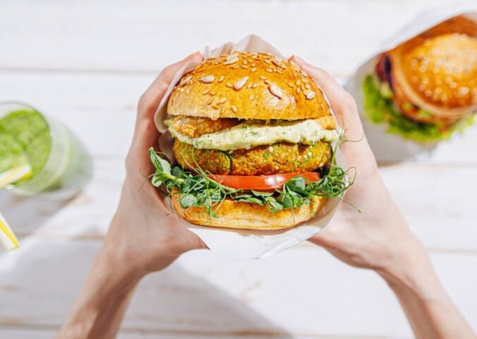Photo shows a woman's hands holding a large vegetarian burger next to a green smoothie and a second sandwich. A new report suggests that ultra-processed plant-based foods are better for heart health than unprocessed animal foods like meat and dairy
