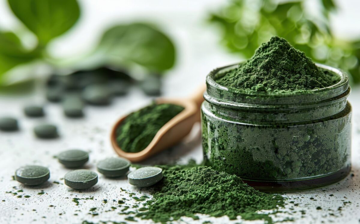 Close-up of green spirulina powder in a jar with tablets and a wooden spoon on a white surface, to showcase the dangers of spirulina