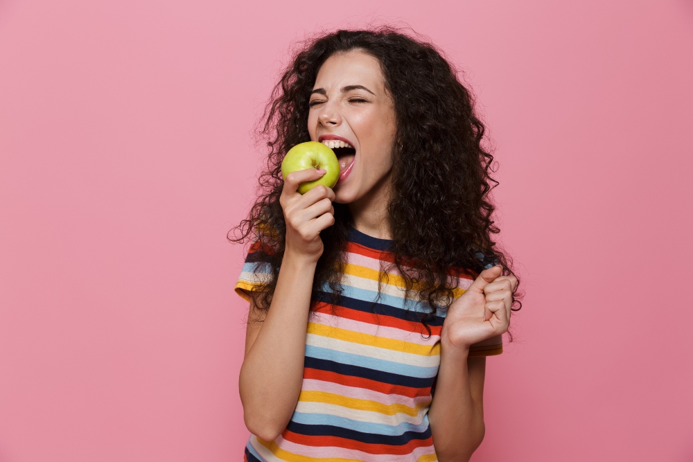 Woman with curly hair eating an apple over a pink background to showcase the dangers of not eating enough fruit in a polluted world