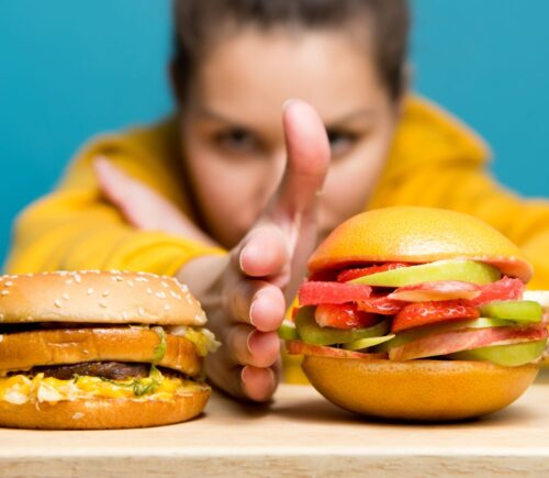 Photo shows a woman holding her hand between two burgers: a traditional beef burger on the left, and a bun filled with sliced fresh fruit on the right