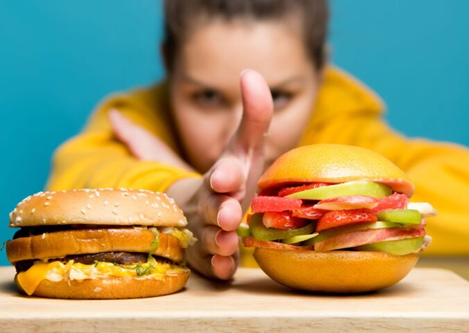 Photo shows a woman holding her hand between two burgers: a traditional beef burger on the left, and a bun filled with sliced fresh fruit on the right