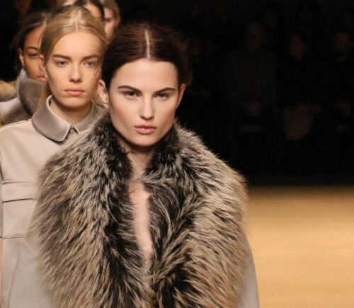 Photo shows a group of models waiting to cross the stage at a fashion event, including one model wearing a large fur coat. New York Fashion Week just announced a fur ban at all events from September 2026