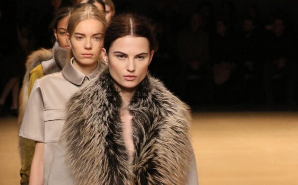 Photo shows a group of models waiting to cross the stage at a fashion event, including one model wearing a large fur coat. New York Fashion Week just announced a fur ban at all events from September 2026