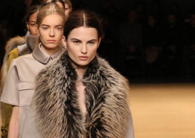 Photo shows a group of models waiting to cross the stage at a fashion event, including one model wearing a large fur coat. New York Fashion Week just announced a fur ban at all events from September 2026
