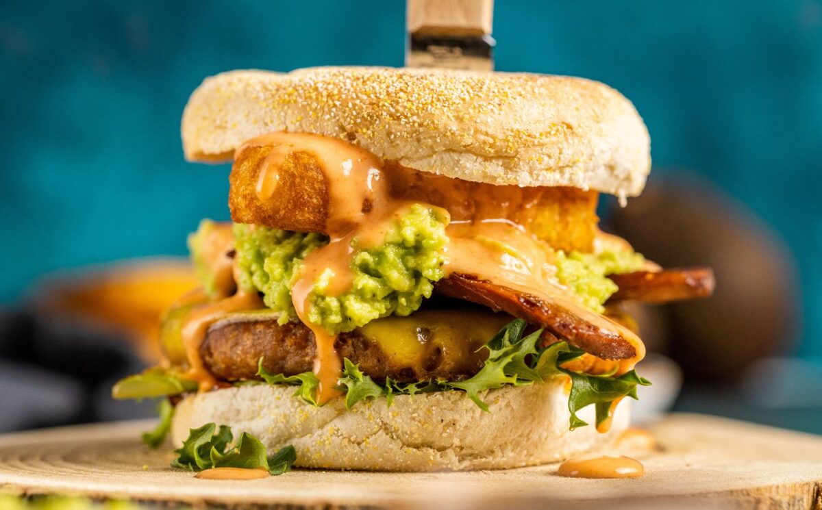 a picture of a stacked vegan breakfast burger made with vegan burger patties, vegan smoky bacon, vegan cheddar, guacamole, and more topped on an English muffin for after NYE