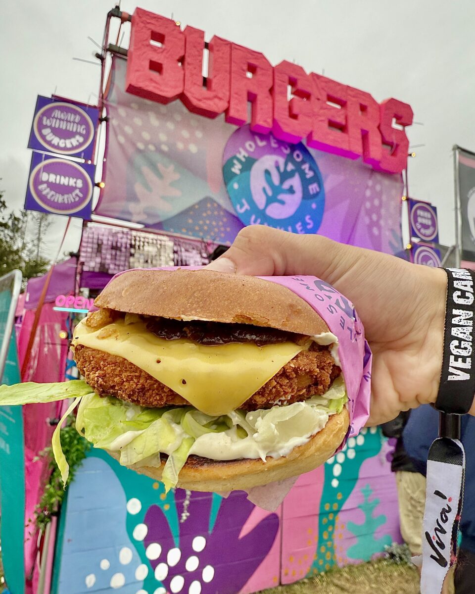 Photo shows someone's hand wearing a Vegan Camp Out wristband and holding up a large cheese and chicken burger in front of a food vendor