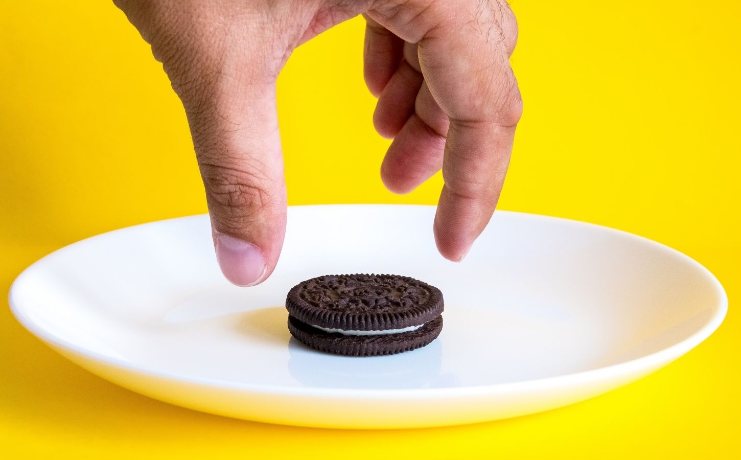 Photo shows someone's hand reaching for an Oreo on a white plate against a bright yellow background. Mondelēz International, the major confectionary multinational behind Oreos, has officially ceased all animal testing