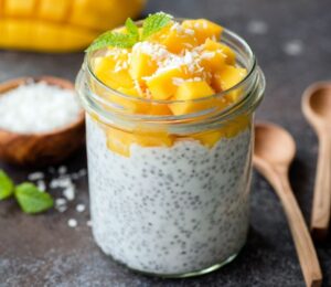 Healthy chia coconut pudding with mango in glass jar and two wooden spoons on a dark background to illustrate the fibermaxxing trend