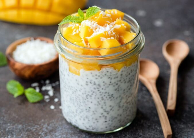 Healthy chia coconut pudding with mango in glass jar and two wooden spoons on a dark background to illustrate the fibermaxxing trend