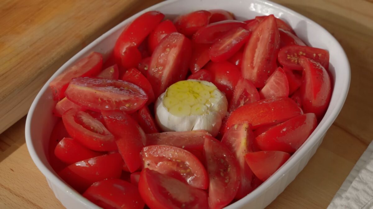 Sliced tomatoes and a head of a garlic in an oven dish: one of the steps to make one of four cozy vegan winter soups.