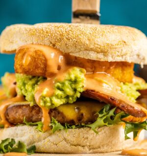 a picture of a stacked vegan breakfast burger made with vegan burger patties, vegan smoky bacon, vegan cheddar, guacamole, and more topped on an English muffin for cozy vegan breakfasts