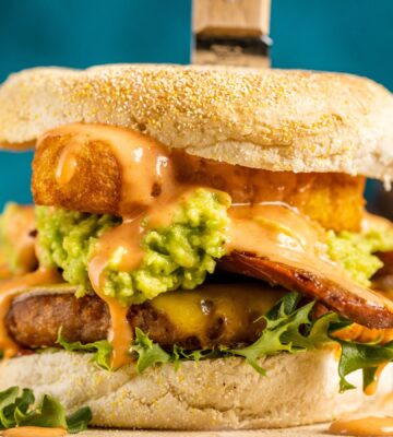 a picture of a stacked vegan breakfast burger made with vegan burger patties, vegan smoky bacon, vegan cheddar, guacamole, and more topped on an English muffin for cozy vegan breakfasts