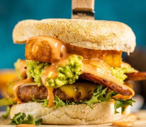 a picture of a stacked vegan breakfast burger made with vegan burger patties, vegan smoky bacon, vegan cheddar, guacamole, and more topped on an English muffin for cozy vegan breakfasts