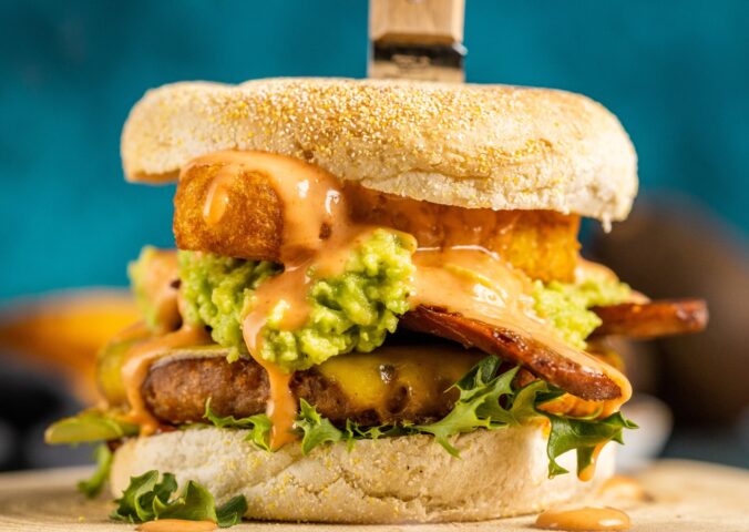 a picture of a stacked vegan breakfast burger made with vegan burger patties, vegan smoky bacon, vegan cheddar, guacamole, and more topped on an English muffin for cozy vegan breakfasts